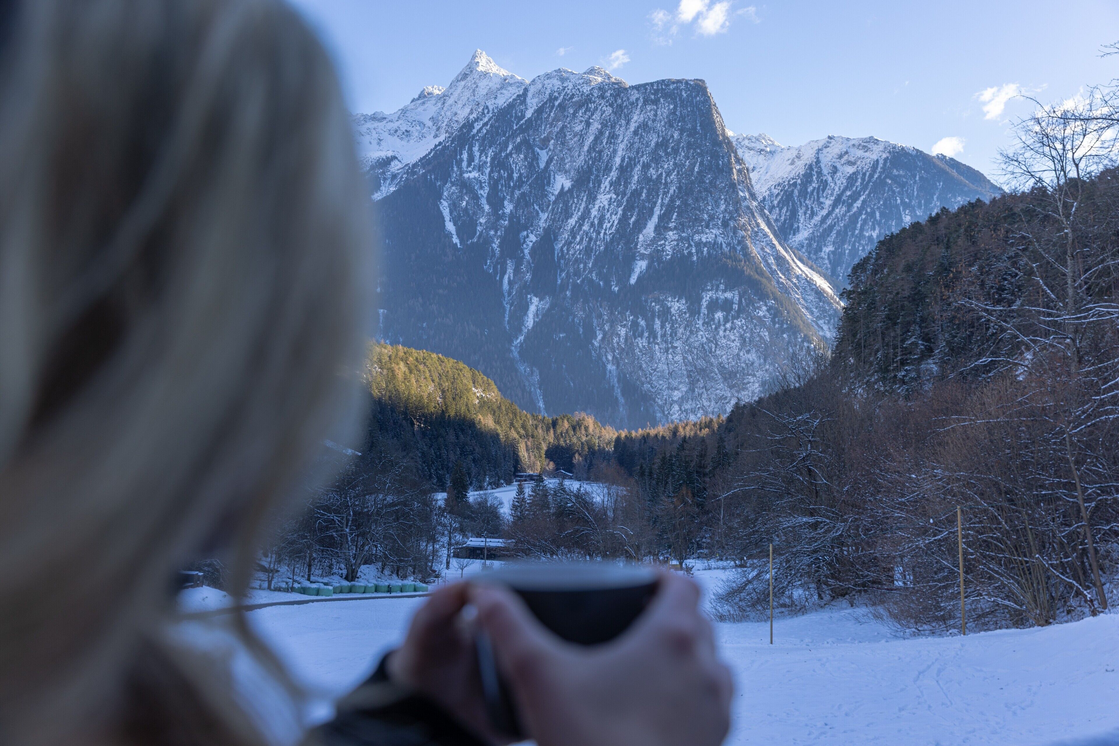 A person holding a cup, overlooking a snowy landscape with mountains and trees.