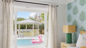 A bedroom with a bed, a nightstand with a lamp, a view of a pool with a pink flamingo float, and a patio area with chairs and a table. - Luxe Villa near Beach, Pool + HotTub, Chef's Kitchen | Woodward Way by AvantStay (Destin)