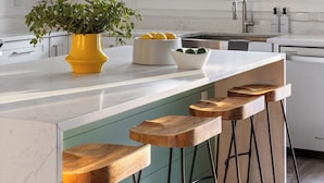 Una cocina moderna con armarios blancos, encimera de mármol, un jarrón amarillo con plantas verdes y taburetes de bar de madera.