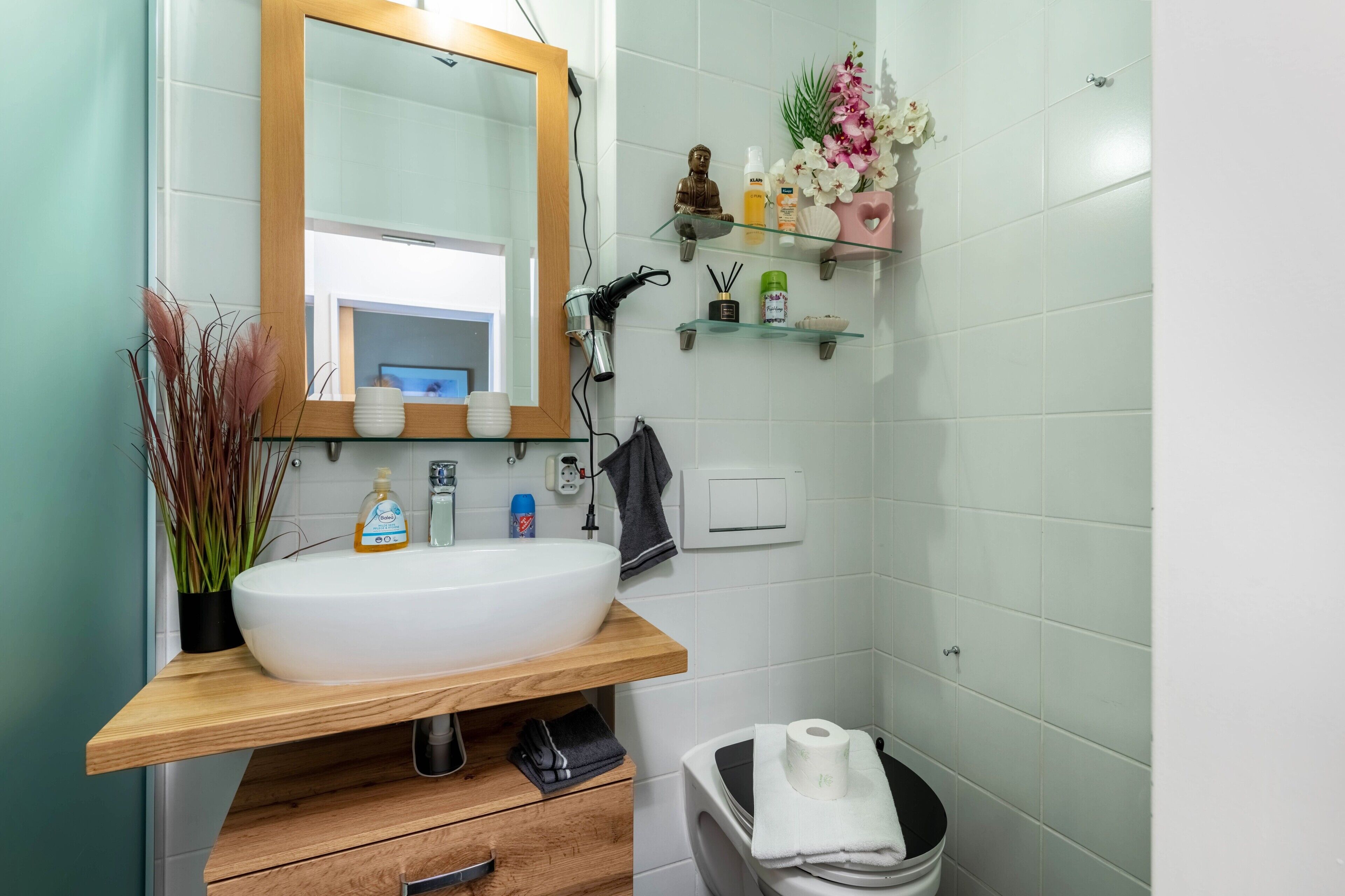Une salle de bain avec un lavabo blanc, un meuble-lavabo en bois, un miroir et une petite étagère contenant des articles de toilette.