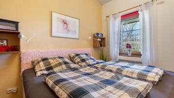 A bedroom with a bed, a bedside table, a wall-mounted lamp, a window with curtains, and a framed picture on the wall.