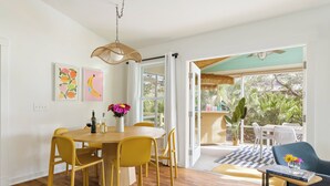 A dining area with a wooden table and yellow chairs, a vase with flowers, and a view of the outdoors through large windows.