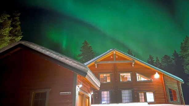 Ein Holzhaus mit Garage und beleuchtetem Eingang, umgeben von Schnee und Bäumen, unter einem Nachthimmel mit grünem Polarlicht.