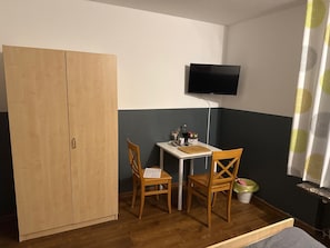 Una habitación con un armario de madera, una pequeña mesa con sillas, un televisor montado en la pared y una cortina estampada.