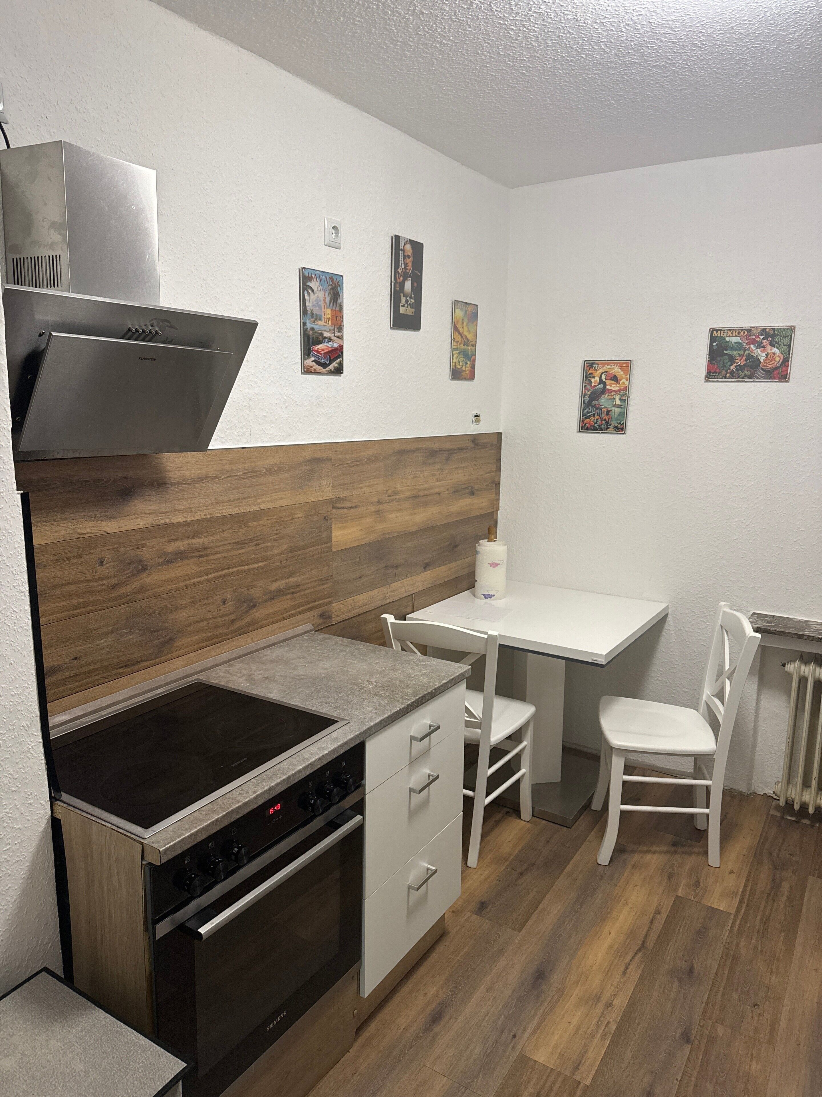 Une cuisine compacte équipée d’une cuisinière, d’un four et d’une hotte intégrés. On y trouve une petite table à manger avec deux chaises. Les murs sont décorés d’affiches sur le thème du voyage.
