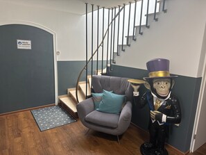 Een trap met een bordje voor handen wassen, een grijze fauteuil met een blauw kussen, en een beeld van een figuur in een hoge hoed.