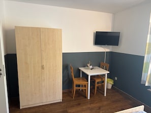 Et værelse med skab, et bord med stole, et vægmonteret fladskærms-tv og et vindue med gardin.