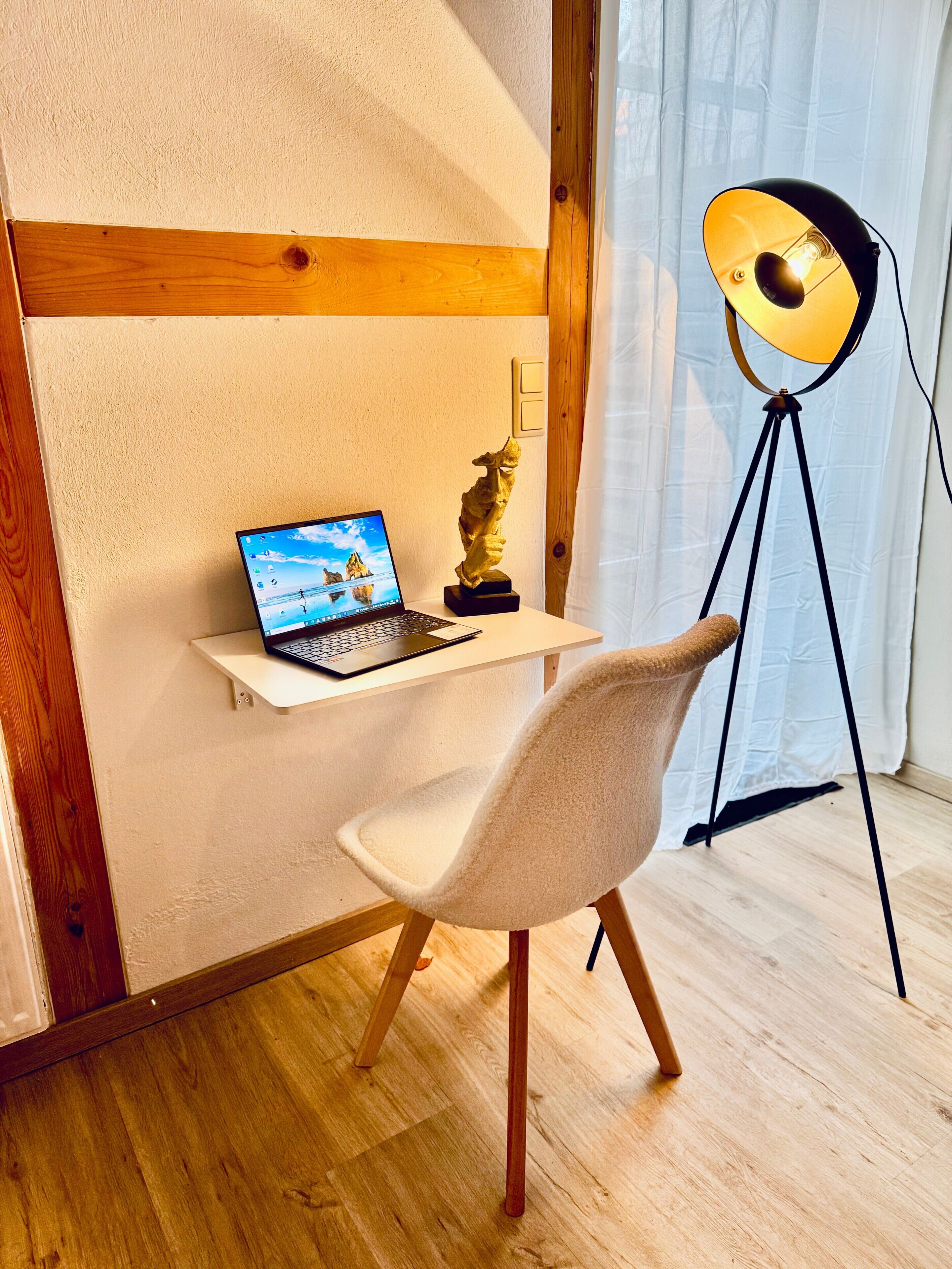 Ein Zimmer mit Schreibtisch, Laptop, einer Skulptur, einem Stuhl und einer Stehlampe.