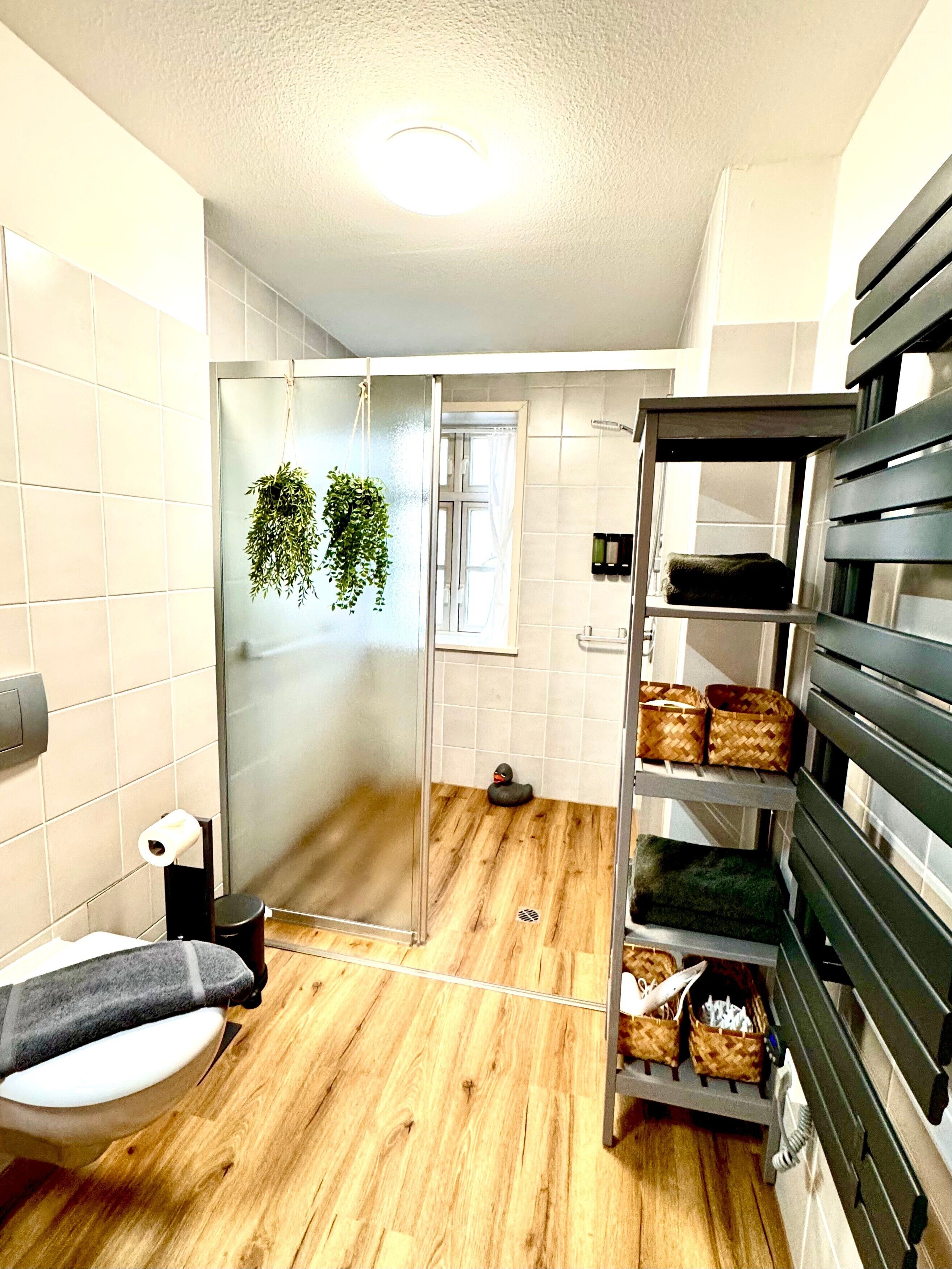 A bathroom with a toilet, a shower, a wooden floor, a towel rack, and a shelf with baskets.