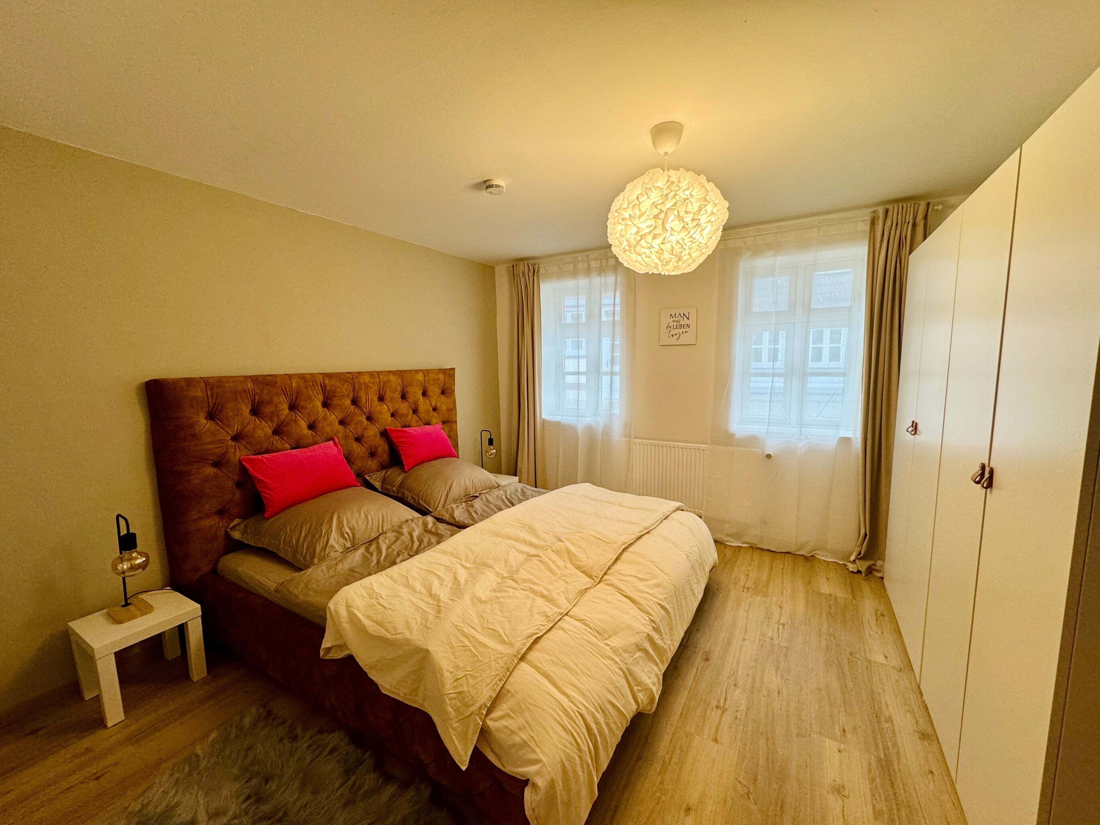 Ein Schlafzimmer mit einer gesteppten Kopfteil, einem Bett mit beiger Bettwäsche, einem kleinen Nachttisch mit Lampe, einem großen Fenster mit Vorhängen und einem Kleiderschrank.