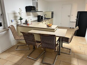 A compact kitchen with a central island, four wicker chairs, a microwave, a coffee maker, and a fruit bowl.