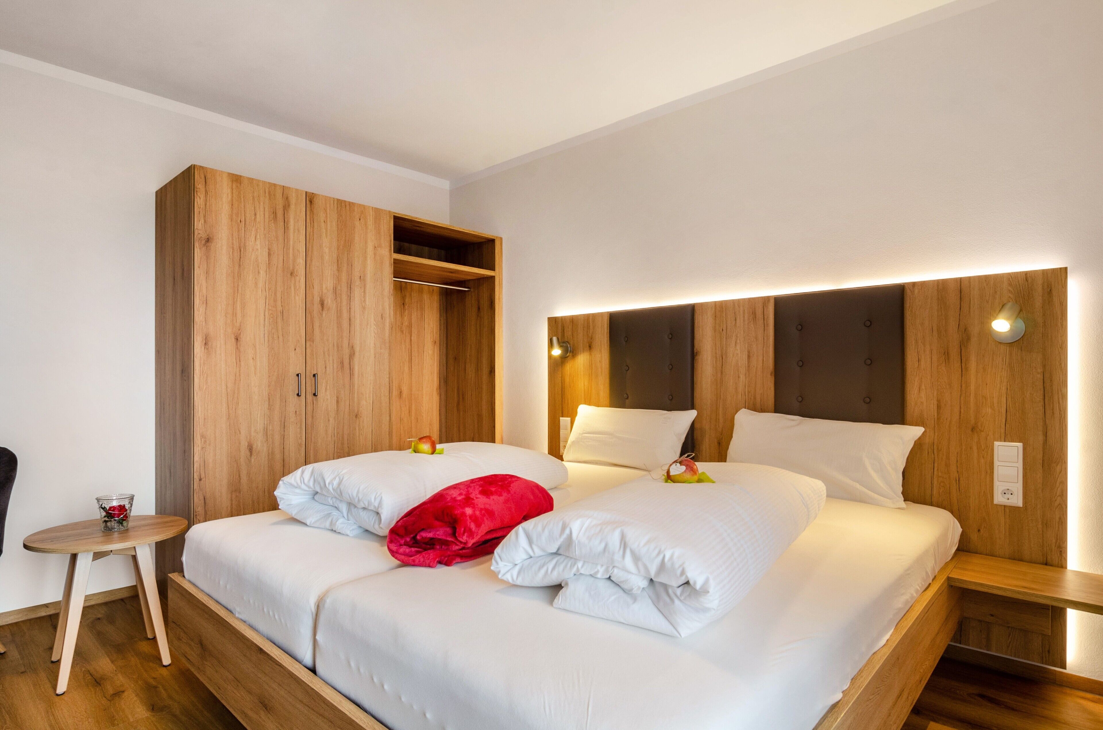 A hotel room with a wooden wardrobe, a bed with white bedding, a small round table, and a bedside table with a lamp.