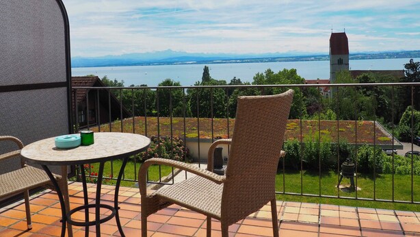 A balcony with wicker furniture, a view of a lake, and a distant tower. - Double Room with Lake View, Balcony and Wi-Fi (Hagnau am Bodensee)