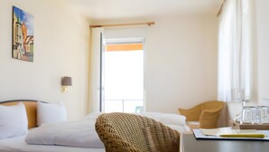 A hotel room with a bed, a wicker chair, a desk with a laptop, and a window with curtains. - Double Room with Lake View, Balcony and Wi-Fi (Hagnau am Bodensee)
