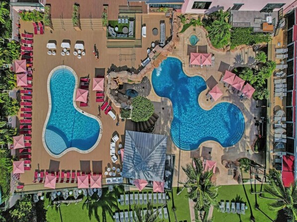 Vista aérea de un complejo turístico con dos piscinas grandes, sillas de descanso y sombrillas.