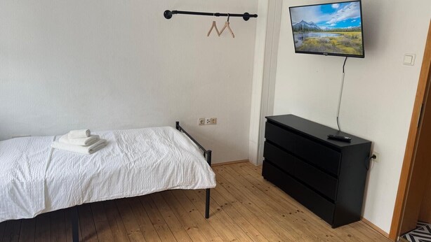 A bedroom with a bed, a black dresser, a TV on the wall, and a wooden floor. - Pfullingen 1 – Accommodation for Workers, Worker House Single Rooms, Wi-Fi (Pfullingen)
