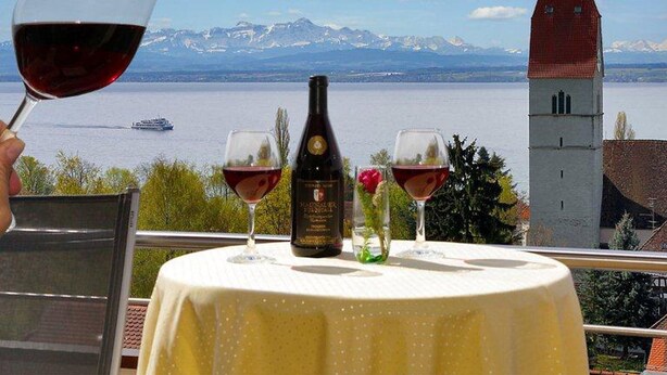 A table with wine glasses and a bottle, overlooking a lake and mountains. - Comfort Room with Side Lake View, Balcony and Wi-Fi (Hagnau am Bodensee)
