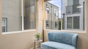 A room with a blue sofa, a small table with a lamp, and a view of neighboring buildings through large windows. - Rooftop Vibes!  Greenlake UW Near Down Town (Seattle)
