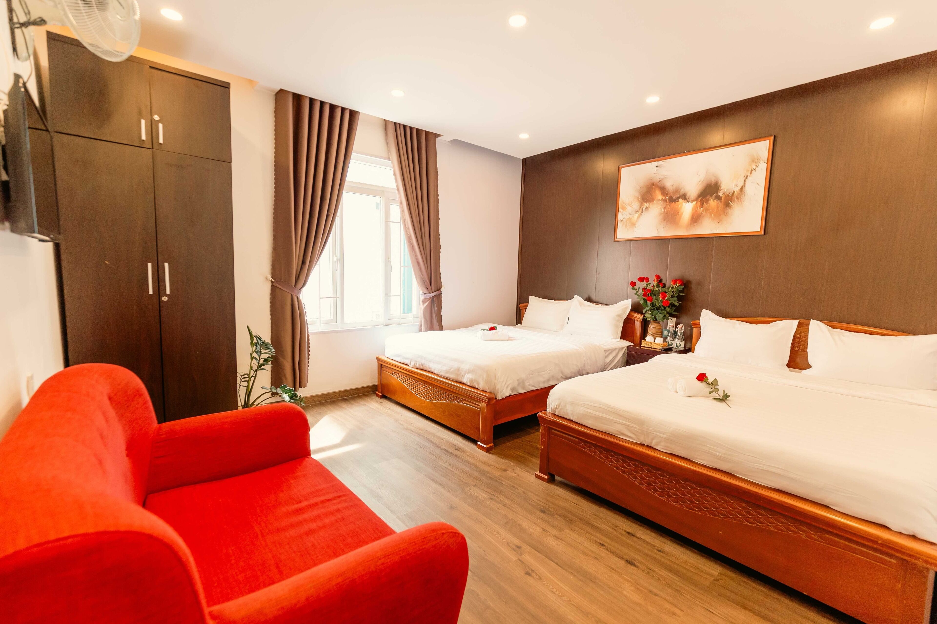 A hotel room with two beds, a red sofa, wooden flooring, a large wardrobe, and a window with curtains.
