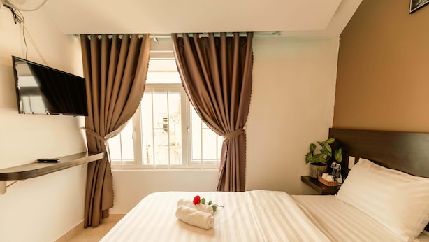 A neatly made bed with white linens and a single red rose, a flat-screen TV mounted on the wall, a bedside table with a plant, and a window with curtains. - An's House  (Xuan Huong - Da Lat)