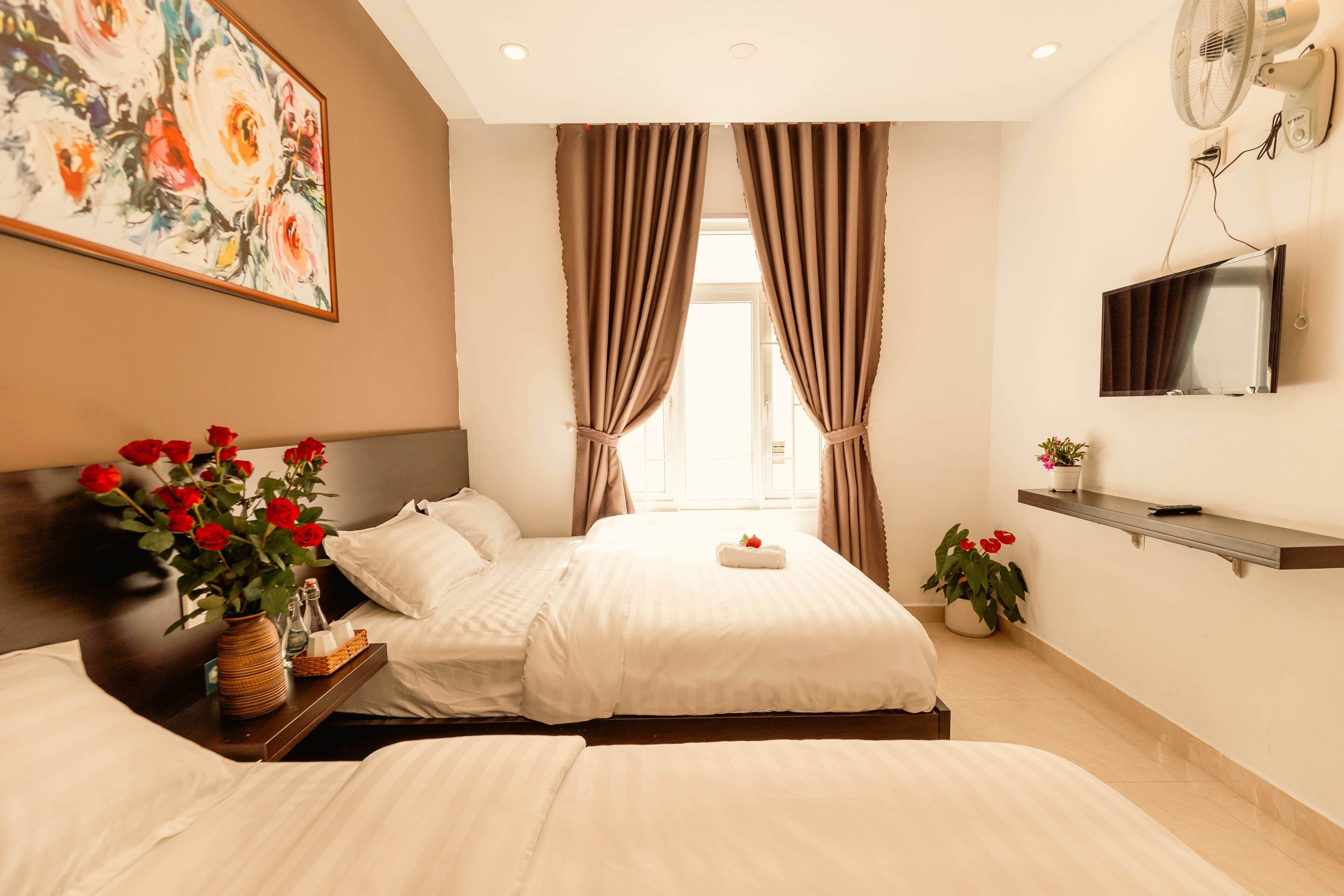 A hotel room with two beds, a TV, a painting, and a vase with red flowers.