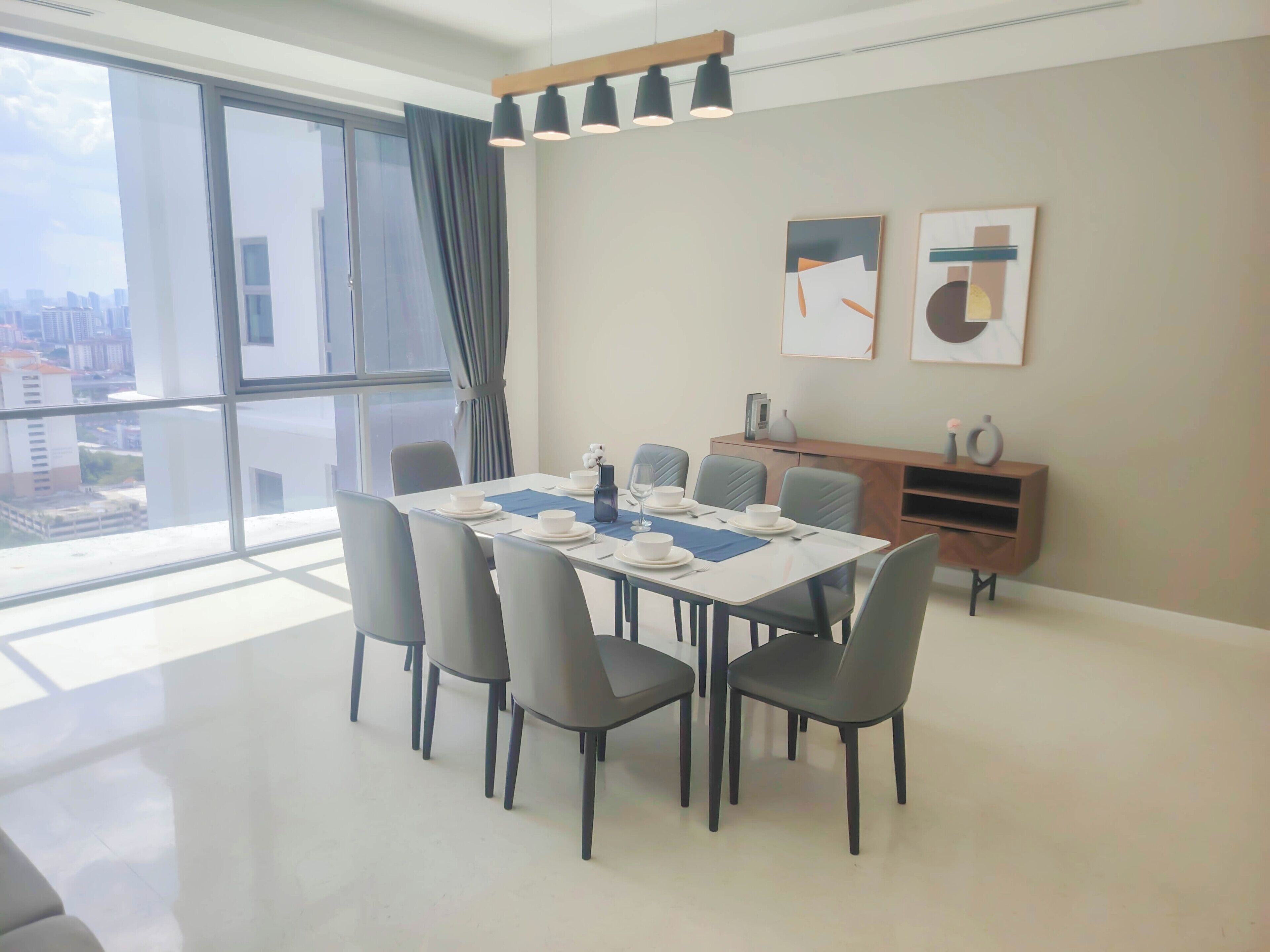 A modern dining area with a large table set for six, surrounded by chairs, in a bright room with large windows and a view of buildings outside.