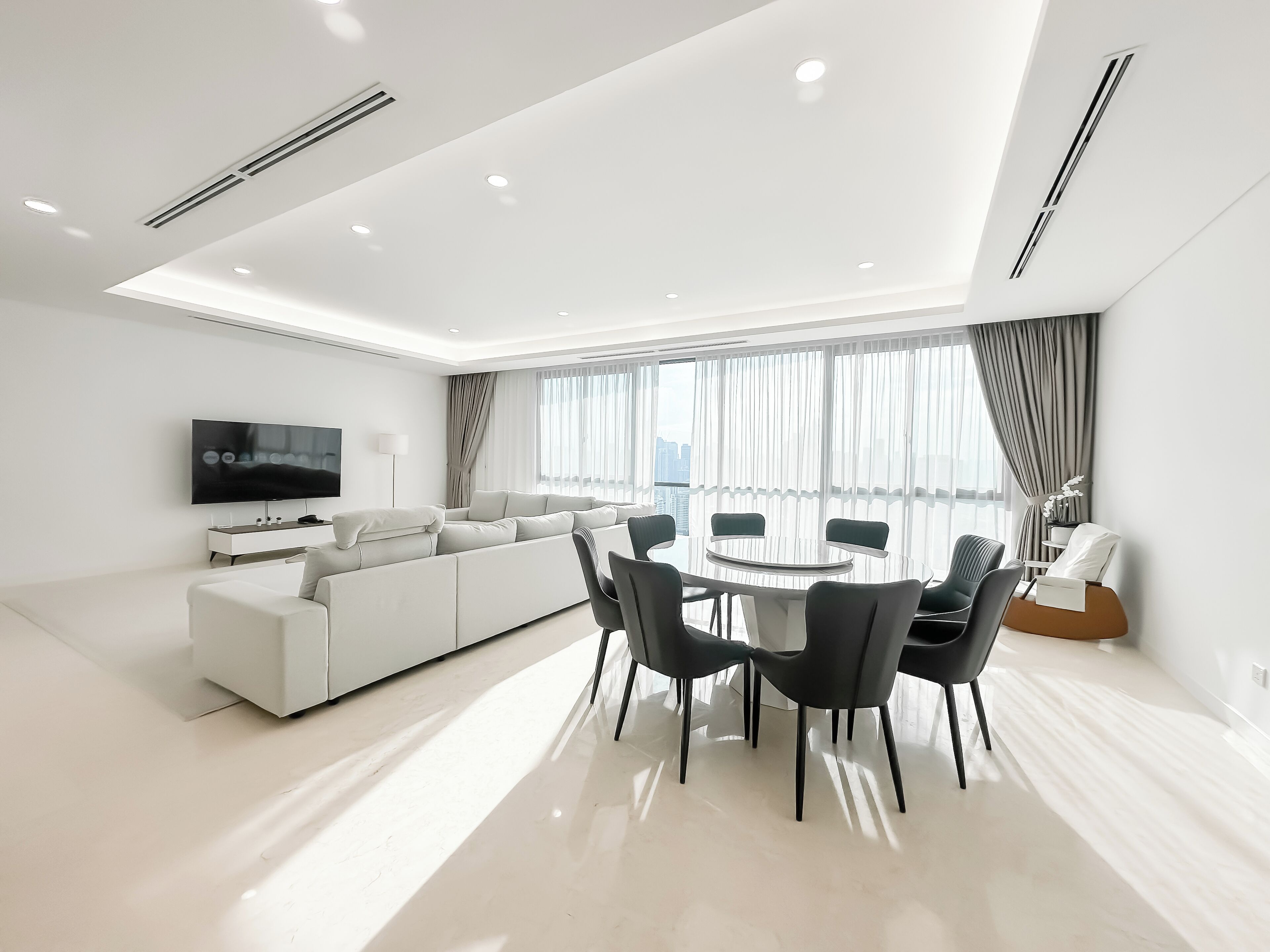 A modern living room with a large white sofa, a dining table with chairs, a flat-screen TV mounted on the wall, and large windows with curtains.