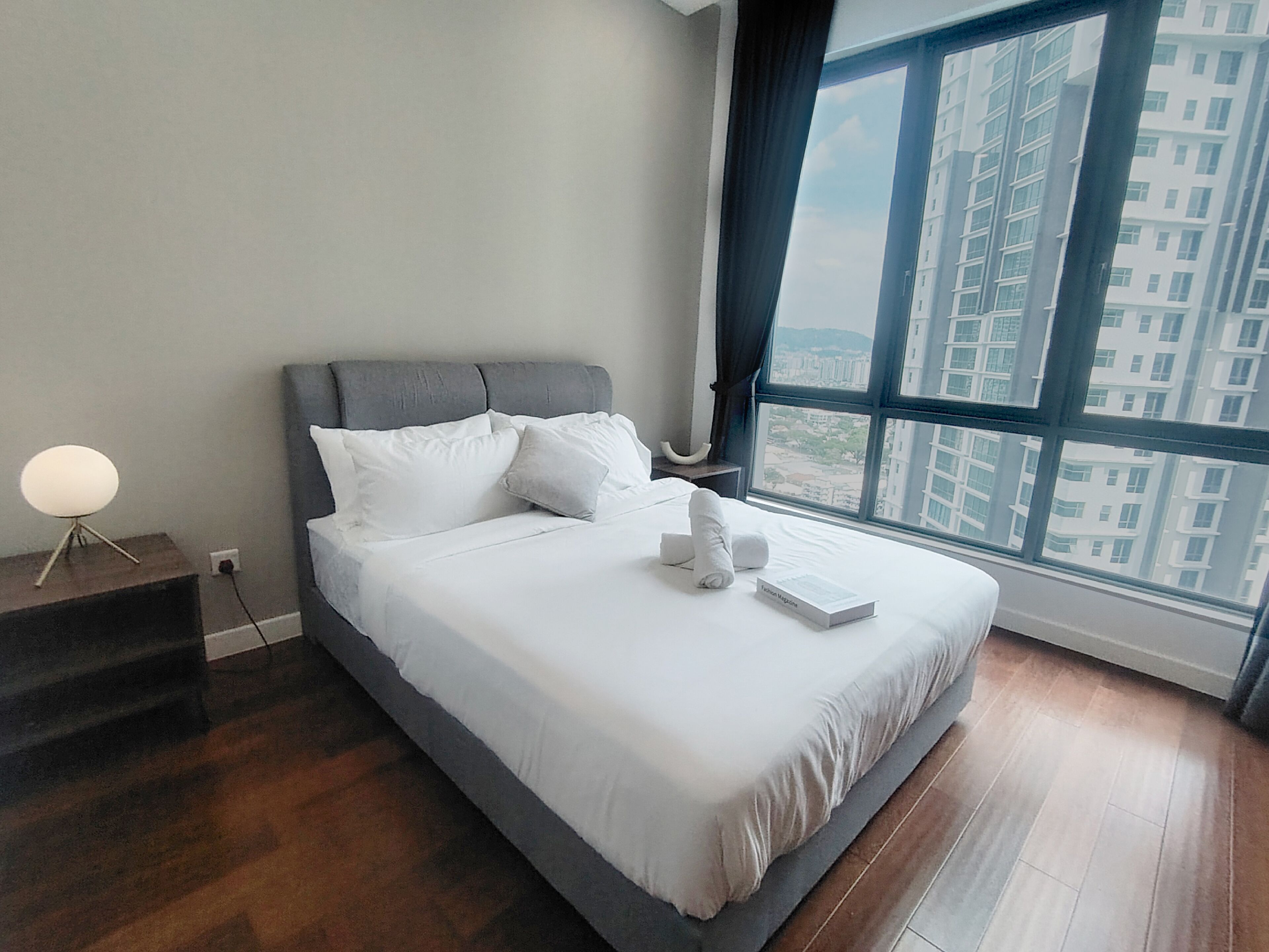 A modern hotel room with a large bed, a bedside lamp, and a view of a cityscape through the window.