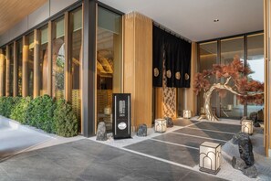 Una entrada de hotel moderna con fachada de vidrio, mostrador de recepción de madera y una escultura de árbol decorativa.
