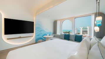 A modern hotel room with a large bed, a flat-screen TV mounted on the wall, a round table, and a seating area with cushions.