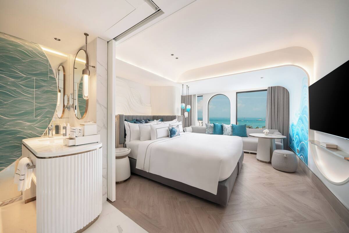 Ein modernes Hotelzimmer mit einem großen Bett, einem Schminktisch mit Spiegel, einem kleinen runden Tisch und Blick auf das Meer.