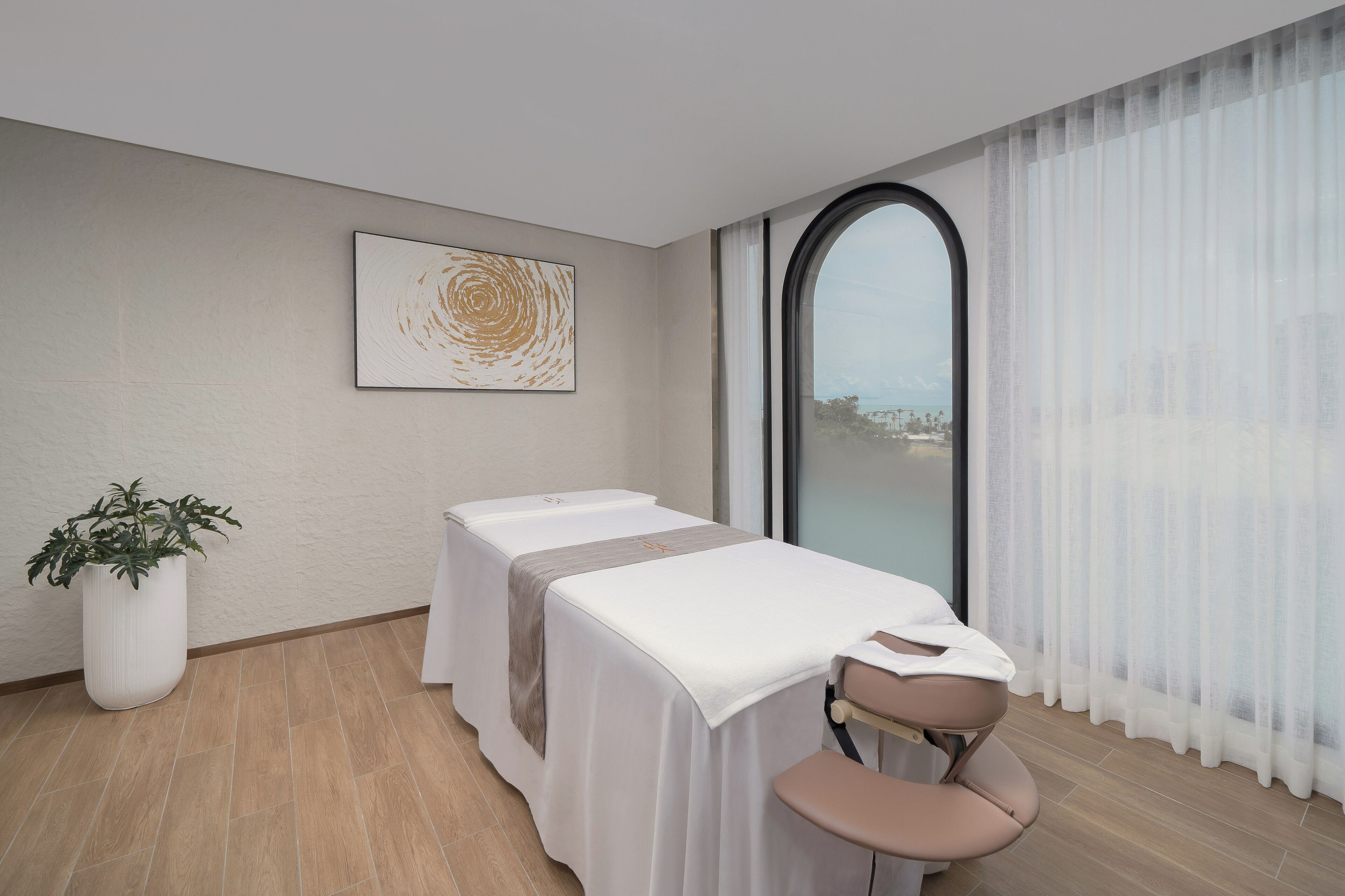 A room with a massage table, a chair, a potted plant, and a large window with a view.