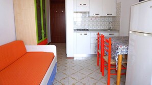 A compact kitchen with white cabinets, a tiled backsplash, a small dining table with chairs, and a bed with an orange mattress.