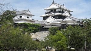 A traditional Japanese castle with multiple levels and a stone base, surrounded by trees and a clear sky. - KKRDOGOYUDUKI (Matsuyama)