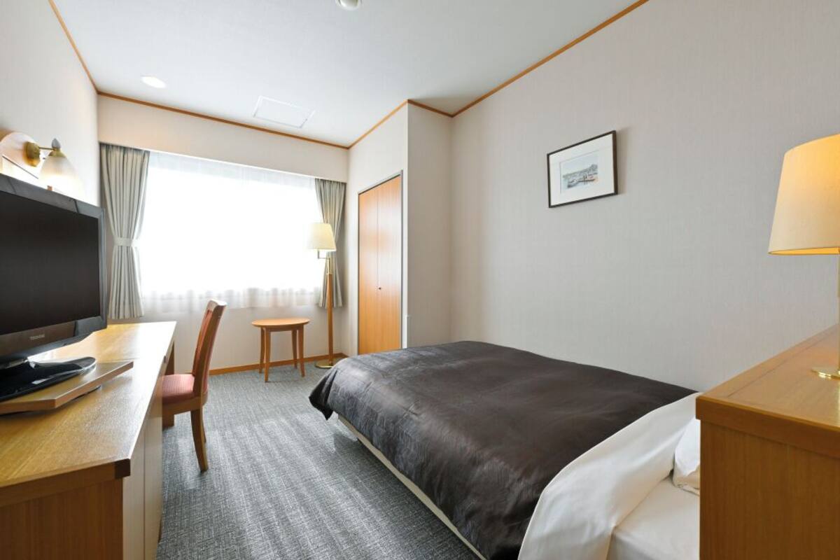Ein Hotelzimmer mit Bett, Schreibtisch, Stuhl, Fernseher und einem Fenster mit Vorhängen.