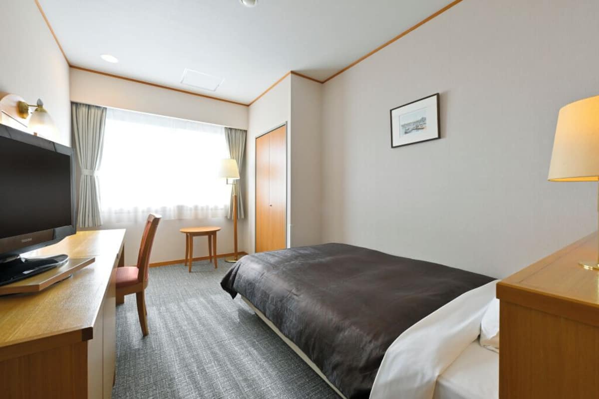 A hotel room with a bed, desk, chair, TV, and a window with curtains.