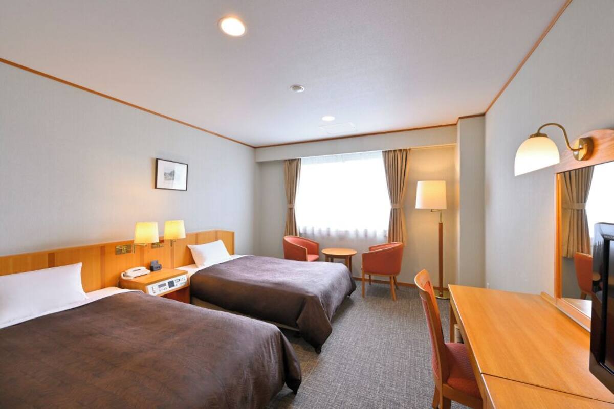 Ein Hotelzimmer mit zwei Betten, einem Schreibtisch, einem Stuhl, einem Fernseher und einem Fenster mit Vorhängen.