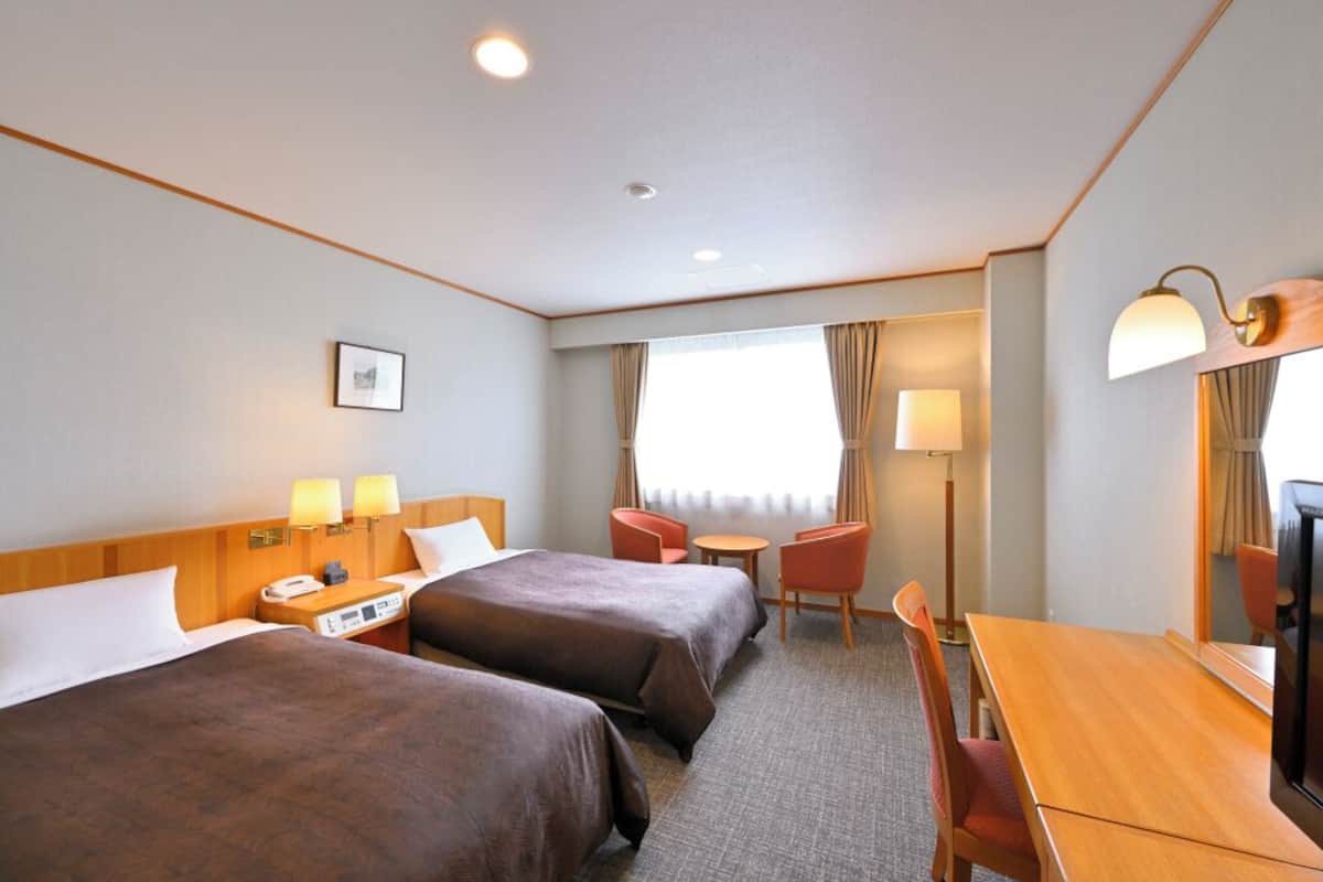 A hotel room with two beds, a desk, a chair, a TV, and a window with curtains.