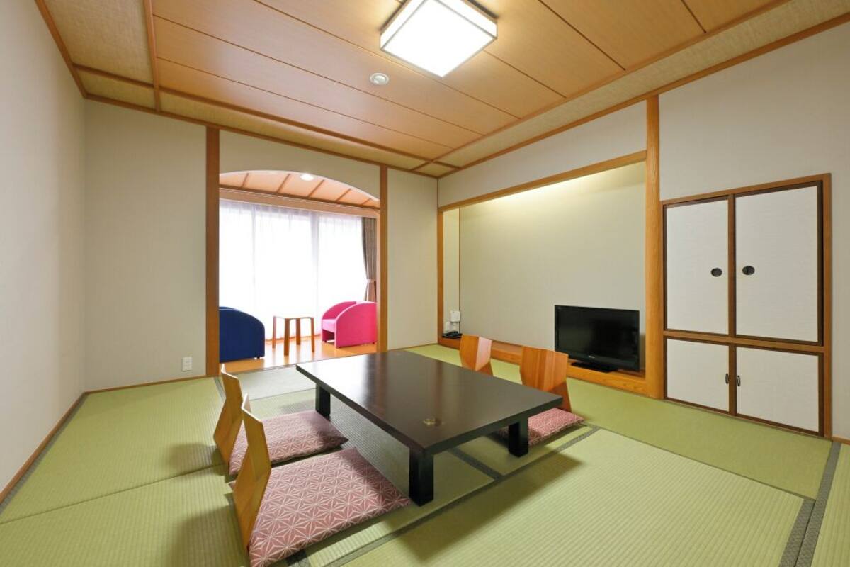 Ein traditioneller japanischer Raum mit Tatami-Bodenbelag, einem niedrigen Tisch, einem Fernseher und Schiebetüren.