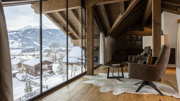 A spacious room with a large window overlooking a snowy landscape, wooden beams on the ceiling, and a modern chair. - Welcoming chalet with balcony (No Deposit) (Ehrwald)