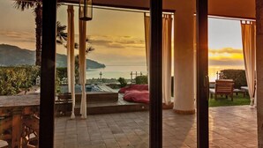 A terrace with a view of the sea and mountains, featuring a wooden table, chairs, and a red bean bag.