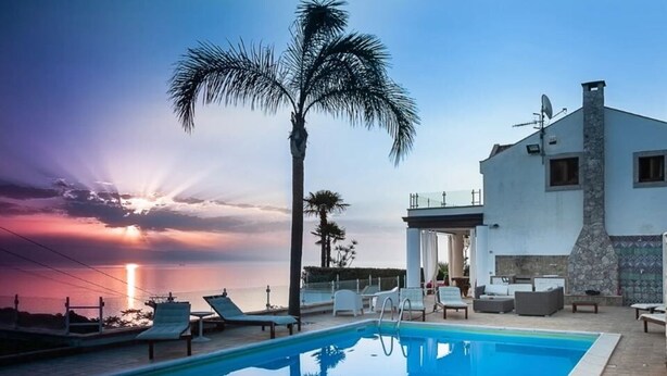 A swimming pool with lounge chairs, a palm tree, and a modern building with a stone facade.