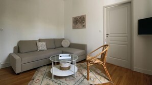 A living room with a sofa, a round coffee table, a wicker chair, a patterned rug, and a door with a flat-screen TV mounted above it.