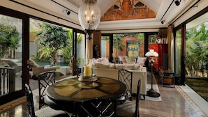 Une pièce spacieuse avec une table à manger ronde, un grand lustre et une vue sur une piscine et une végétation luxuriante à l’extérieur.