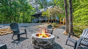 Ein Steinkaminfeuer, umgeben von Adirondack-Stühlen, einer Holzterrasse und einem Haus mit überdachter Veranda, eingebettet in einen Wald.
