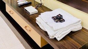 A hotel room with a wooden vanity, a flat-screen TV mounted on the wall, a water dispenser, a remote control, a digital scale, and a stack of white towels. - Hotel Panchavati Palace (Khandwa)