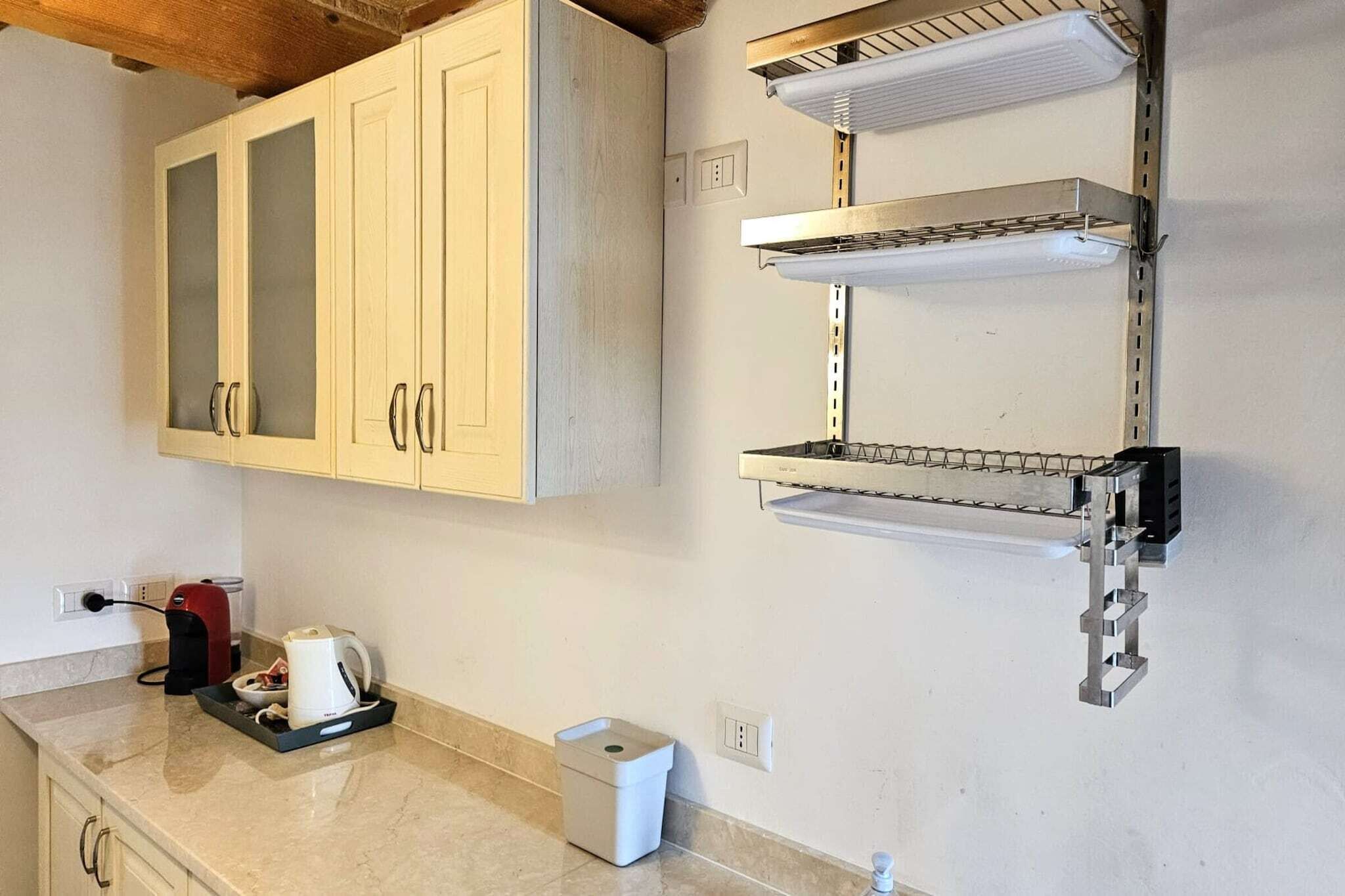 A kitchen with light wooden cabinets, a stainless steel range hood, a kettle, and a trash bin.