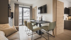 A modern living room with a sofa, a dining table with chairs, a flat-screen TV mounted on the wall, and a large window with curtains. - Apartment in Kirchberg near Schaterzlift (Kirchberg in Tirol)
