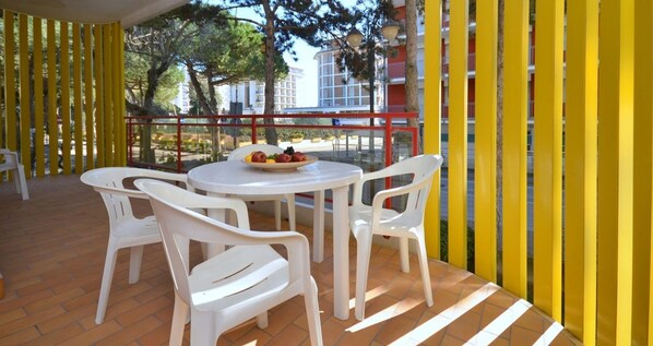 Een overdekt terras met witte plastic stoelen en een tafel, gele verticale zonwering, en een uitzicht op gebouwen in de verte.
