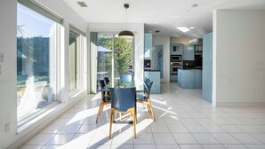 Cozinha moderna com ilha central, grandes janelas e área de refeições com mesa e cadeiras de vidro.